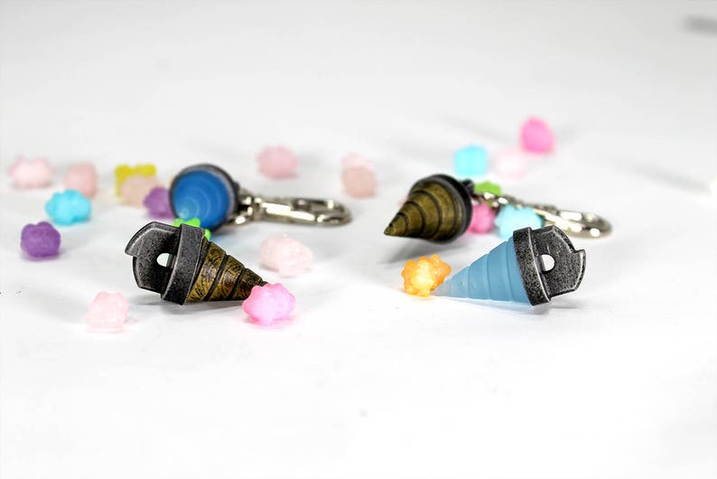May include: Three different drill bit keychains, one blue, one brown, and one gray, with silver metal accents. The keychains are scattered on a white surface with small colorful candies.