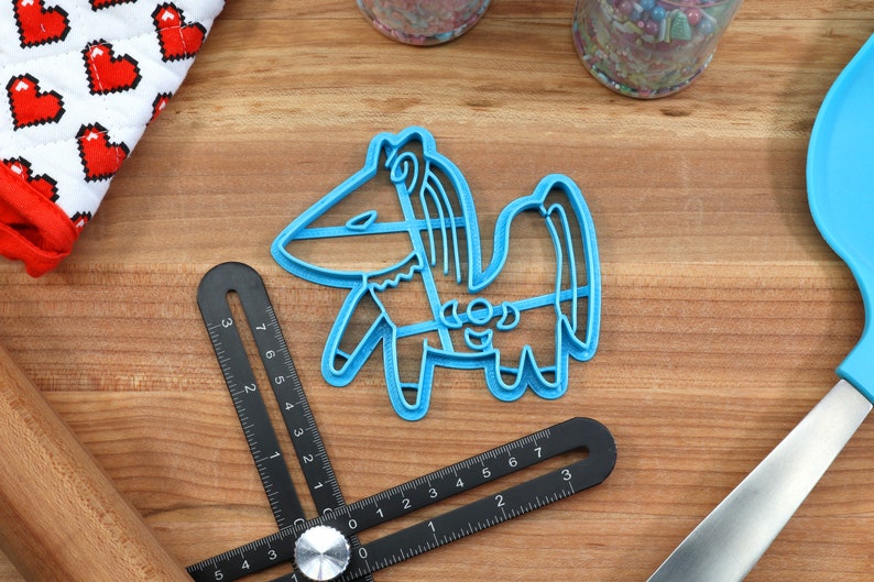 Puede incluir: Un cortador de galletas de pl&aacute;stico azul con forma de caballo de dibujos animados con melena y cola. El cortador est&aacute; sobre una superficie de madera con una regla y un rodillo.