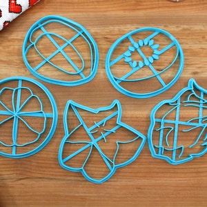 May include: Six blue plastic cookie cutters shaped like different fruits and vegetables. The cutters are on a wooden surface.
