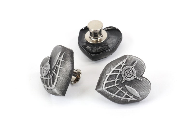 K&ouml;nnte beinhalten: Drei silberne herzf&ouml;rmige Metallkn&ouml;pfe mit einem dekorativen Design. Die Kn&ouml;pfe haben eine strukturierte Oberfl&auml;che und ein erhabenes Design aus einem Netz und einem Ziel.