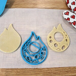 May include: Three cookie cutters in the shape of a flame. The cutters are made of blue plastic and are on a white parchment paper surface.