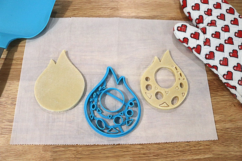 May include: Three cookie cutters in the shape of a flame. The cutters are made of blue plastic and are on a white parchment paper surface.
