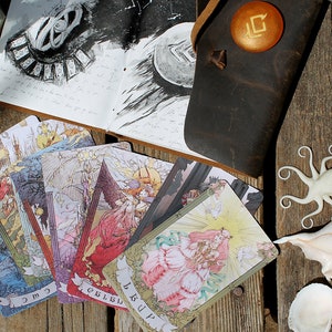 May include: A set of tarot cards with colorful illustrations on a wooden surface. The cards are spread out in a fan shape, with the top card showing a woman in a pink dress. There is a leather journal with a drawing of a moon and a black and white sketch of a clock face. A white octopus figurine and a seashell are also visible.