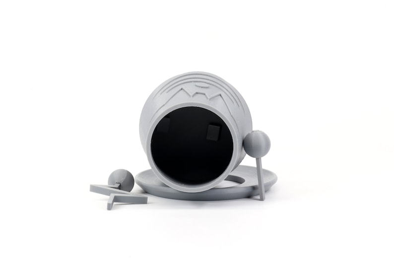 Pode incluir: Um recipiente cinzento, impresso em 3D, com a parte superior aberta, uma tampa e duas pequenas al&ccedil;as redondas. O recipiente est&aacute; sobre uma superf&iacute;cie branca.