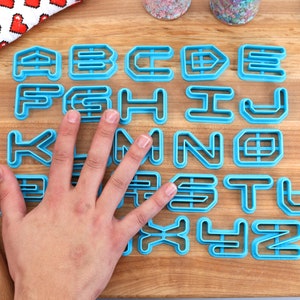 May include: A set of blue plastic alphabet cookie cutters on a wooden surface. The cutters are arranged in alphabetical order.