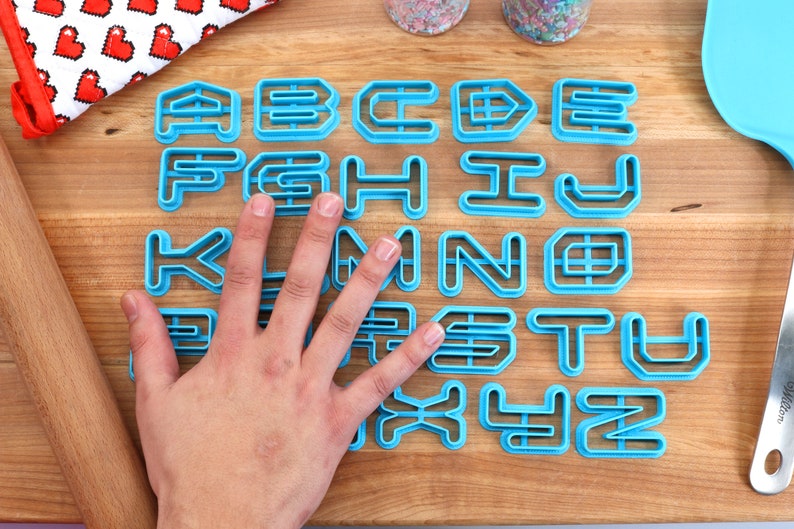 May include: A set of blue plastic alphabet cookie cutters on a wooden surface. The cutters are arranged in alphabetical order.