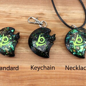 May include: Three black resin pendants with a green and gold glitter design and a green symbol. The pendants are shown on a wooden surface with the words "Standard", "Keychain", and "Necklace" underneath each pendant.