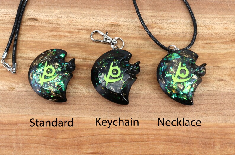 May include: Three black resin pendants with a green and gold glitter design and a green symbol. The pendants are shown on a wooden surface with the words "Standard", "Keychain", and "Necklace" underneath each pendant.