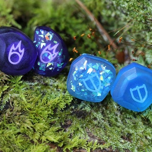 May include: Four resin dice with iridescent glitter embedded in the resin. Two dice are purple with a flame symbol, and two dice are blue with a shield symbol.