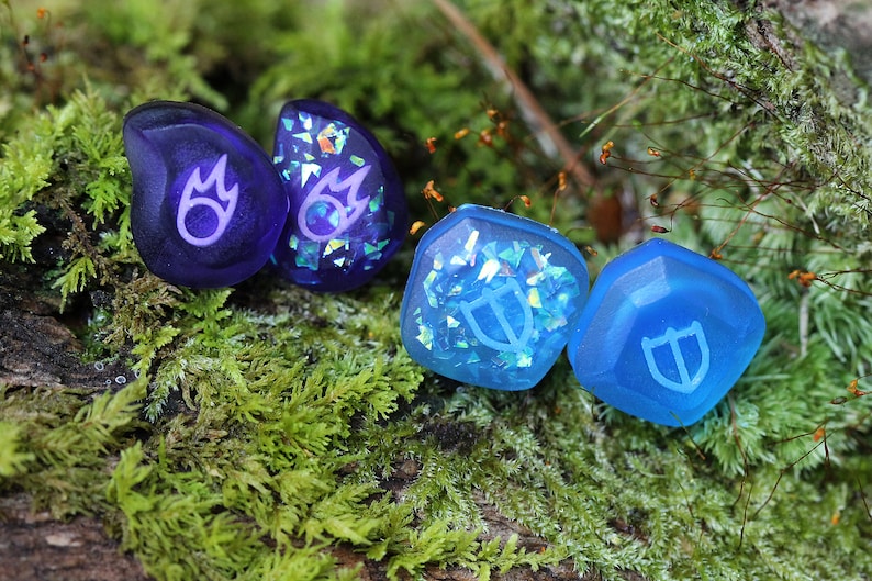 May include: Four resin dice with iridescent glitter embedded in the resin. Two dice are purple with a flame symbol, and two dice are blue with a shield symbol.