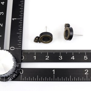 Puede incluir: Un par de pendientes de bot&oacute;n negros y dorados con un peque&ntilde;o dise&ntilde;o redondo. Los pendientes se muestran sobre un fondo blanco con una regla negra.