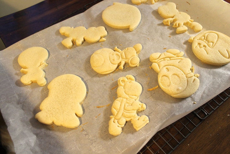 May include: A sheet of parchment paper with various cookie shapes on it. The cookies are unbaked and have a light brown color. The shapes include a circle, a star, a mushroom, a snowman, a penguin, and a turtle.