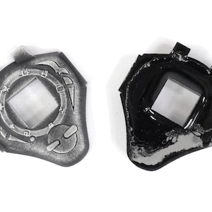 May include: Two silver-colored plastic toy keychains shaped like a shield with a square window in the center. One keychain is detailed with a circular design and the other is plain black.