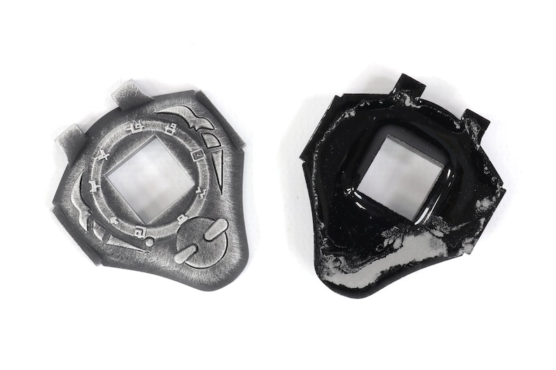May include: Two silver-colored plastic toy keychains shaped like a shield with a square window in the center. One keychain is detailed with a circular design and the other is plain black.