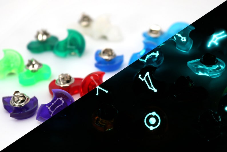May include: A set of 10 glow-in-the-dark constellation pins. The pins are shaped like crescent moons and feature different constellations. The pins are shown in both daylight and dark conditions, highlighting their glow-in-the-dark feature.