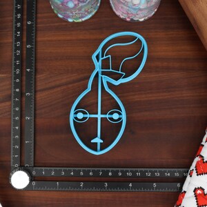 May include: A light blue plastic cookie cutter shaped like a cartoon character with a large head and a small body. The character has two large eyes, a small nose, and a mouth.