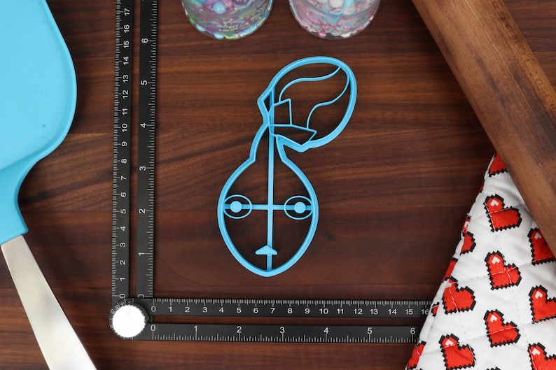 May include: A light blue plastic cookie cutter shaped like a cartoon character with a large head and a small body. The character has two large eyes, a small nose, and a mouth.