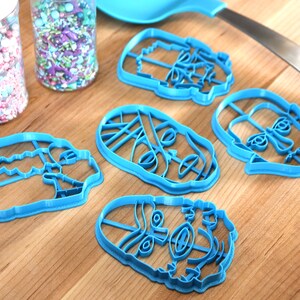 May include: A collection of blue plastic cookie cutters in various face shapes, alongside jars of colorful sprinkles and a blue spatula. The cutters are arranged on a wooden surface, suggesting a baking theme.