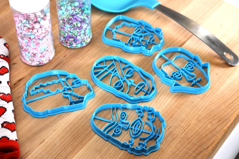 May include: A collection of blue plastic cookie cutters in various face shapes, alongside jars of colorful sprinkles and a blue spatula. The cutters are arranged on a wooden surface, suggesting a baking theme.