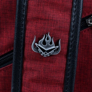 May include: A red canvas backpack with a black zipper and a silver metal pin featuring a stylized flame design.