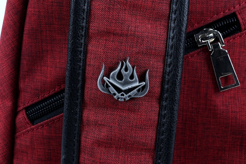 May include: A red canvas backpack with a black zipper and a silver metal pin featuring a stylized flame design.