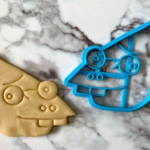 K&ouml;nnte beinhalten: Ein blauer Plastik-Ausstecher in Form einer Comicfigur mit einem &uuml;berraschten Ausdruck und ein Cookie, der in Form des Ausstechers gebacken wurde.
