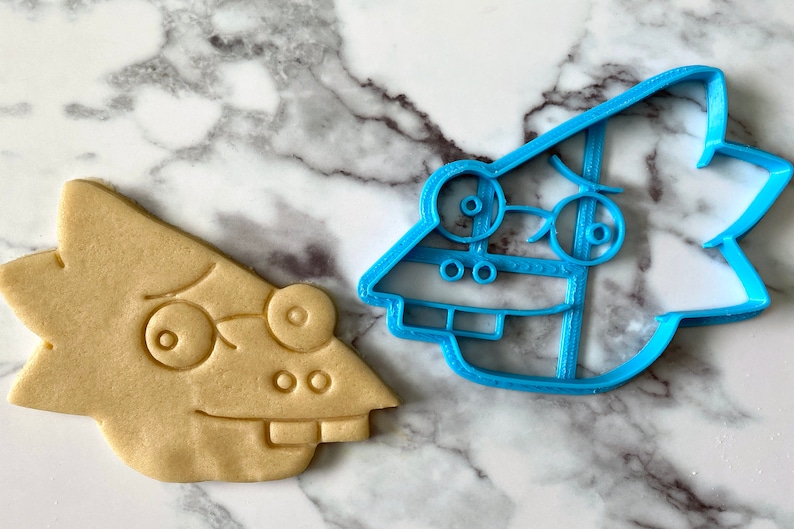 K&ouml;nnte beinhalten: Ein blauer Plastik-Ausstecher in Form einer Comicfigur mit einem &uuml;berraschten Ausdruck und ein Cookie, der in Form des Ausstechers gebacken wurde.