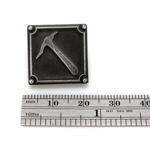 K&ouml;nnte beinhalten: Eine silberfarbene Metallnadel mit quadratischer Form und einem erhabenen Bild einer Spitzhacke. Die Nadel ist etwa 2 cm breit.