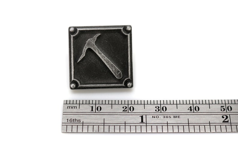 K&ouml;nnte beinhalten: Eine silberfarbene Metallnadel mit quadratischer Form und einem erhabenen Bild einer Spitzhacke. Die Nadel ist etwa 2 cm breit.