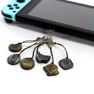 Może przedstawiać: Zestaw sześciu srebrnych i złotych brelok&oacute;w z r&oacute;żnymi symbolami. Breloki są przymocowane do konsoli Nintendo Switch.