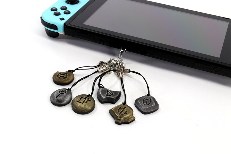 Może przedstawiać: Zestaw sześciu srebrnych i złotych brelok&oacute;w z r&oacute;żnymi symbolami. Breloki są przymocowane do konsoli Nintendo Switch.