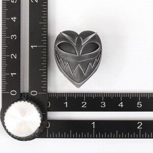 May include: A silver heart-shaped pin with a black and white grinning face design.
