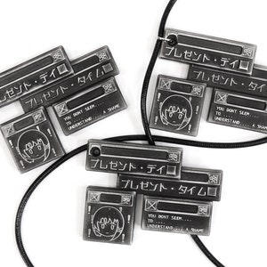 May include: Two black and silver necklaces with pendants featuring the Japanese text "プレゼント・デイ" and "プレゼント・タイム" along with a cartoon face. The necklaces are on a white background.