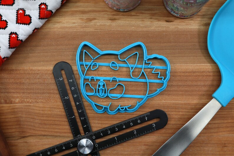 May include: Blue plastic cookie cutter shaped like a cartoon dog with a striped pattern. The cutter is on a wooden surface.