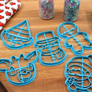 Op de afbeelding: Een set van zes blauwe plastic kerstkoekjesvormen, waaronder een sneeuwpop, rendier, elf, peperkoekman en kerstman. De vormen liggen op een houten oppervlak.