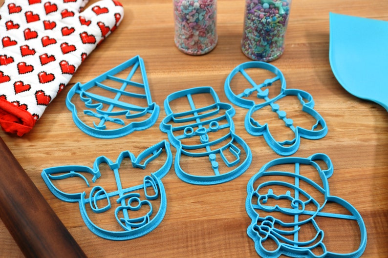 Op de afbeelding: Een set van zes blauwe plastic kerstkoekjesvormen, waaronder een sneeuwpop, rendier, elf, peperkoekman en kerstman. De vormen liggen op een houten oppervlak.