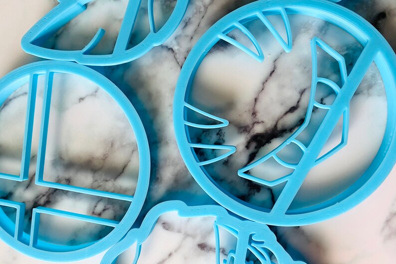 May include: A set of five blue plastic cookie cutters in various shapes, including a circle with a cross inside, a circle with a crescent moon shape inside, and a leaf shape. The cutters are on a white marble surface.