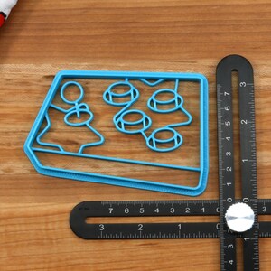 May include: A blue plastic cookie cutter shaped like a retro arcade game with four buttons. The cookie cutter is about 3 inches wide.