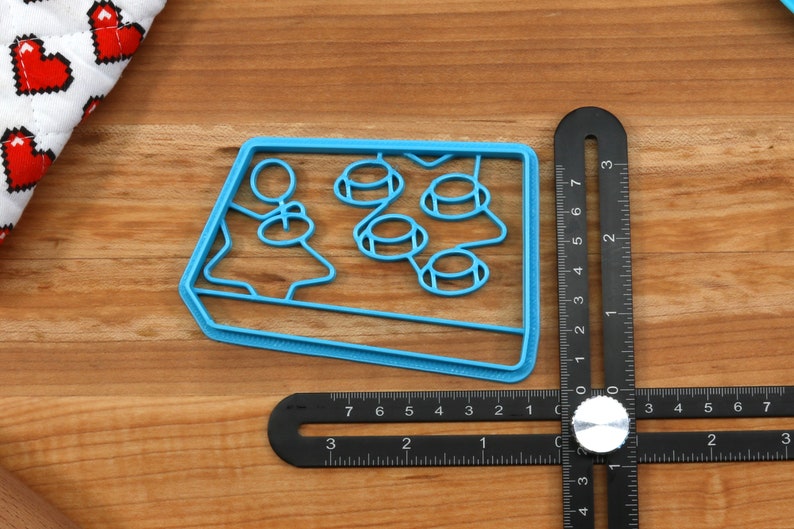 May include: A blue plastic cookie cutter shaped like a retro arcade game with four buttons. The cookie cutter is about 3 inches wide.
