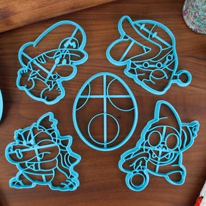 Op de afbeelding: Vijf blauwe plastic koekjesvormen in de vorm van videogamekarakters. De vormen liggen op een bruin houten oppervlak.