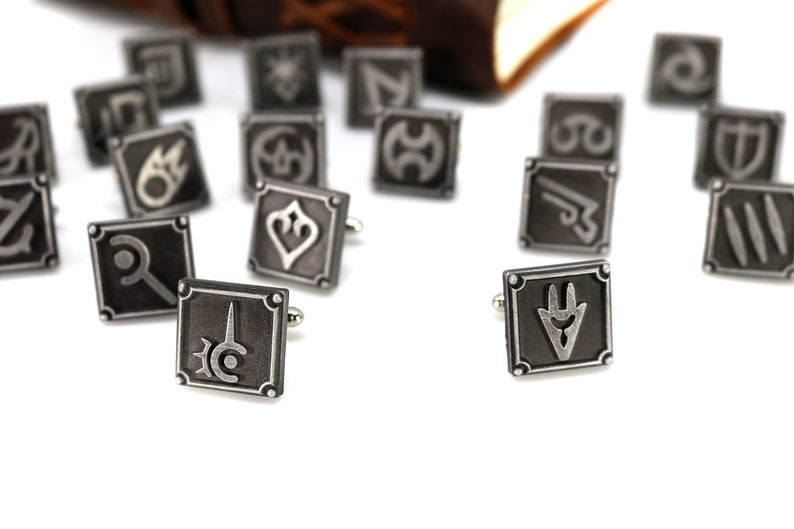 May include: A set of silver cufflinks featuring various symbols, including a sword, a shield, and a crescent moon. The cufflinks are scattered on a white surface.