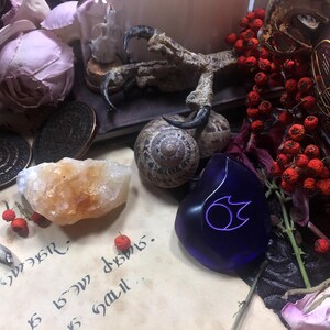 Op de afbeelding: Een blauwe glazen amulet met een gestileerd vlamontwerp, omringd door gedroogde bessen, een slakkenhuis en een kristal.