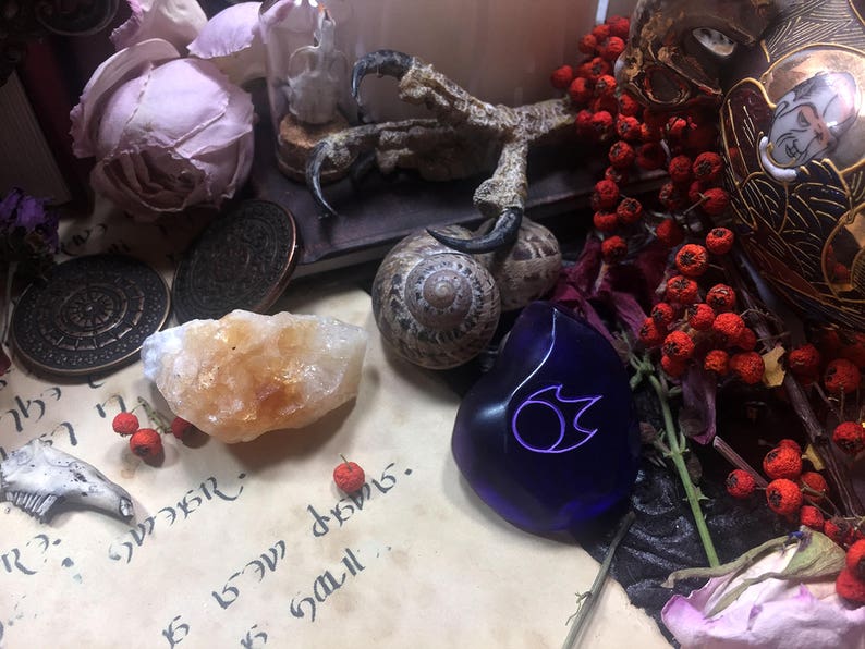 Op de afbeelding: Een blauwe glazen amulet met een gestileerd vlamontwerp, omringd door gedroogde bessen, een slakkenhuis en een kristal.