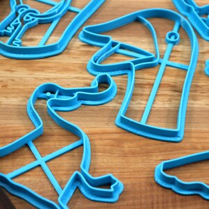 May include: A set of blue plastic dinosaur cookie cutters on a wooden surface. The cutters are shaped like different dinosaurs, including a T-Rex, a Triceratops, and a Stegosaurus.