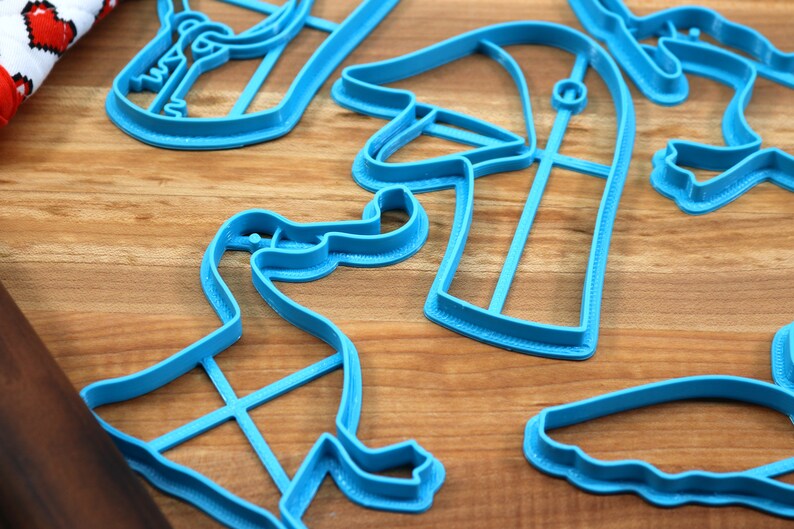 May include: A set of blue plastic dinosaur cookie cutters on a wooden surface. The cutters are shaped like different dinosaurs, including a T-Rex, a Triceratops, and a Stegosaurus.