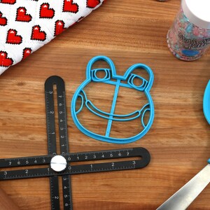 May include: A blue plastic frog-shaped cookie cutter on a wooden surface with a black ruler, a rolling pin, and a blue spatula.