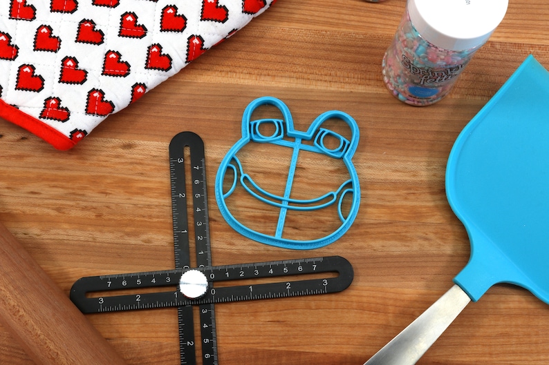 May include: A blue plastic frog-shaped cookie cutter on a wooden surface with a black ruler, a rolling pin, and a blue spatula.
