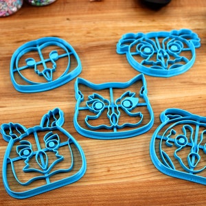 May include: Five blue plastic cookie cutters shaped like animal faces. The cutters are on a wooden surface. The animals are an owl, a koala, a cat, a deer, and a pug.