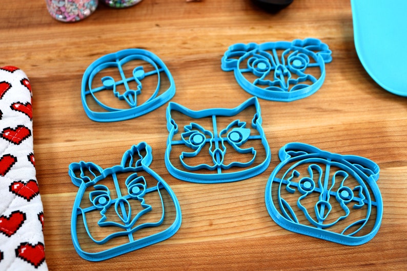 May include: Five blue plastic cookie cutters shaped like animal faces. The cutters are on a wooden surface. The animals are an owl, a koala, a cat, a deer, and a pug.
