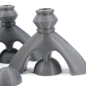 May include: Three gray, 3D printed candle holders with a unique, geometric design. Each holder has a round base and three curved arms that meet at the top to form a triangular opening. The holders are made of a smooth, matte material.